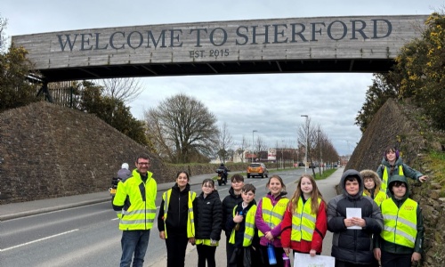 Group of students standing in hi-vis underneath the 'Welcome to Sherford' sign. 
